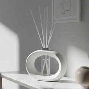 White oval ceramic diffuser with reeds on a marble table in a bright, minimalist room, with a small beige vase nearby.