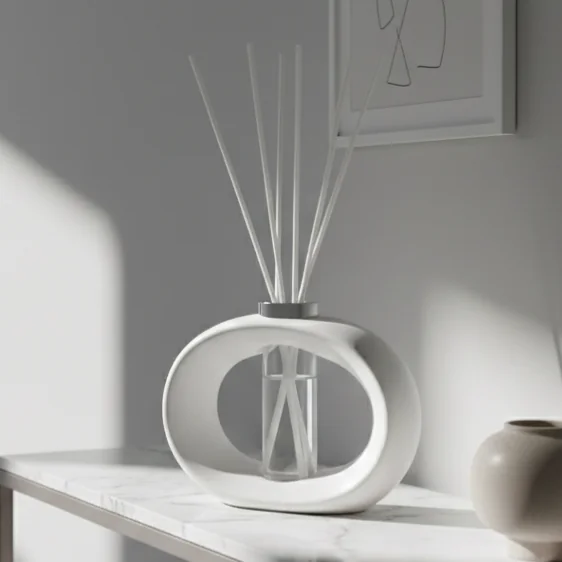 White oval ceramic diffuser with reeds on a marble table in a bright, minimalist room, with a small beige vase nearby.